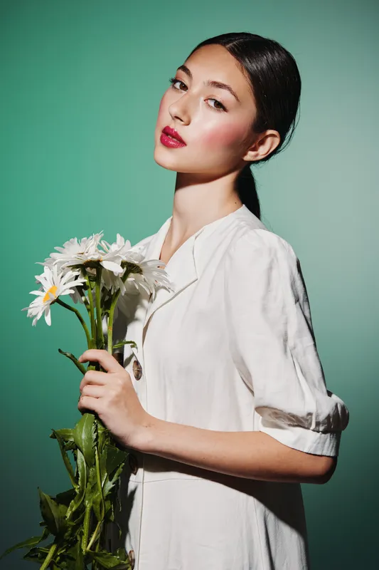 Young woman with luminous skin and vivid makeup holds a bouquet of daisies. She wears a relaxed white shirt against a green backdrop, showcasing beauty, fashion, and creative styling.