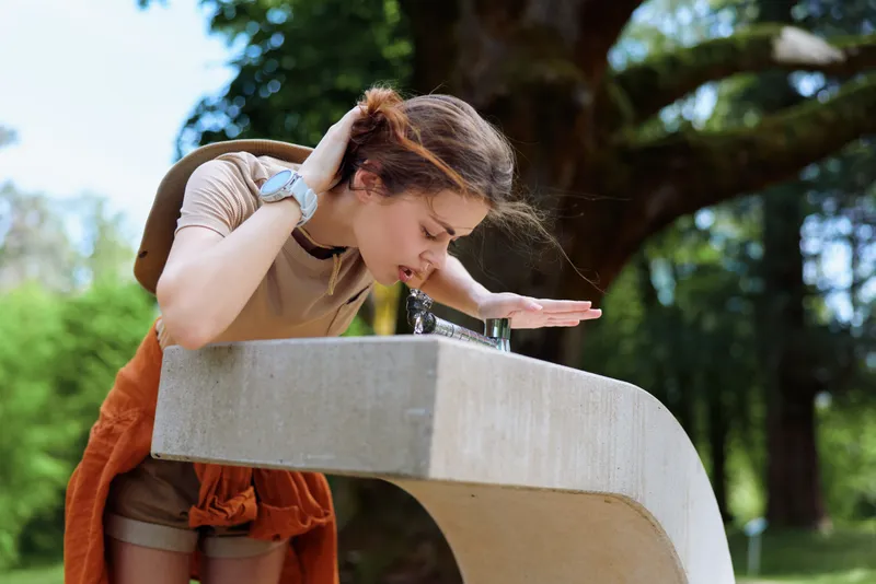 Woman drinking from a concrete park fountain outdoors with backpack and summer shorts, seeking hydration and refreshment during a casual nature outing, candid portrait capturing sunlight and movement.