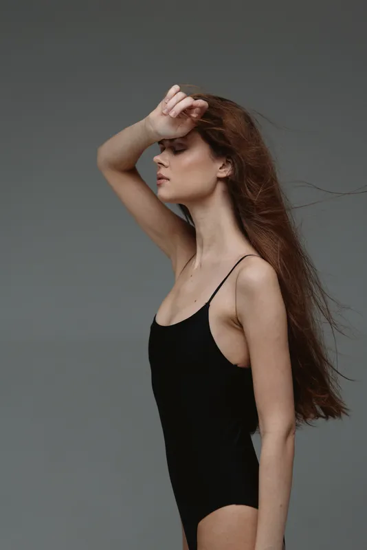 Profile of a slender woman in black bodysuit with long hair posing against a gray background, showing elegance and calm expression.