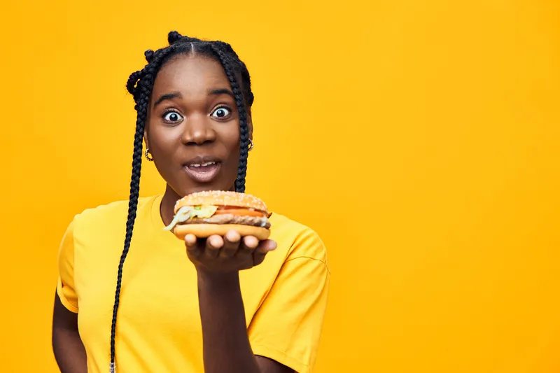 Happy Black woman holding a delicious burger against a vibrant yellow background, showcasing excitement and joy in a casual summer outfit