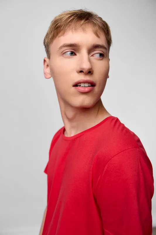 Young man in a red shirt turns his head to the side against a neutral studio backdrop, expressing curiosity, style, and casual confidence in soft lighting.