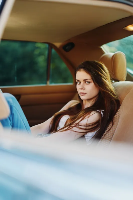 A contemplative young woman with long brown hair relaxing in a car, wearing a casual white tank top and jeans, capturing the essence of summer freedom.
