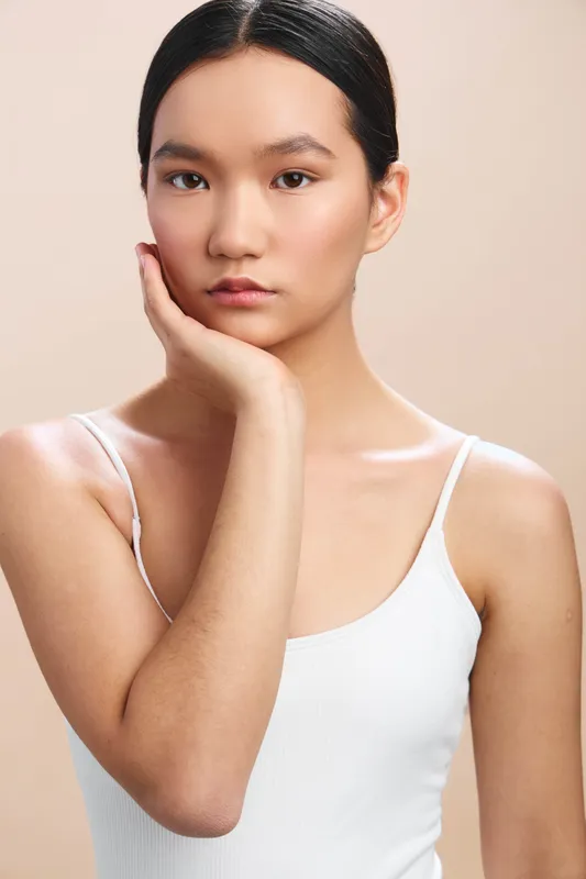 Asian woman in a simple pose rests her chin on her hand, wearing a light tank top. The soft neutral backdrop emphasizes calm confidence and poised, contemporary elegance.