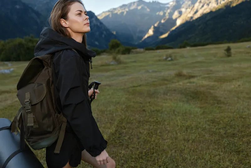 An adventurous woman with a backpack stands in a scenic mountain meadow, looking ahead with determination and curiosity, enjoying fresh air and expansive views, while the valley and distant peaks
