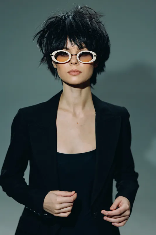 Stylish woman wearing white sunglasses and black blazer with confident expression in studio lighting