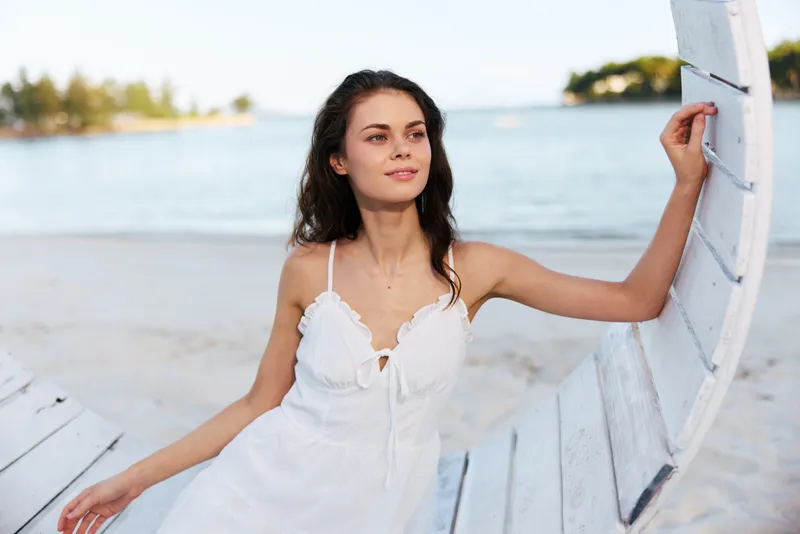 A young woman in a white dress stands on a sandy beach, leaning against a curved wooden frame near the shore, gazing softly into the distance.