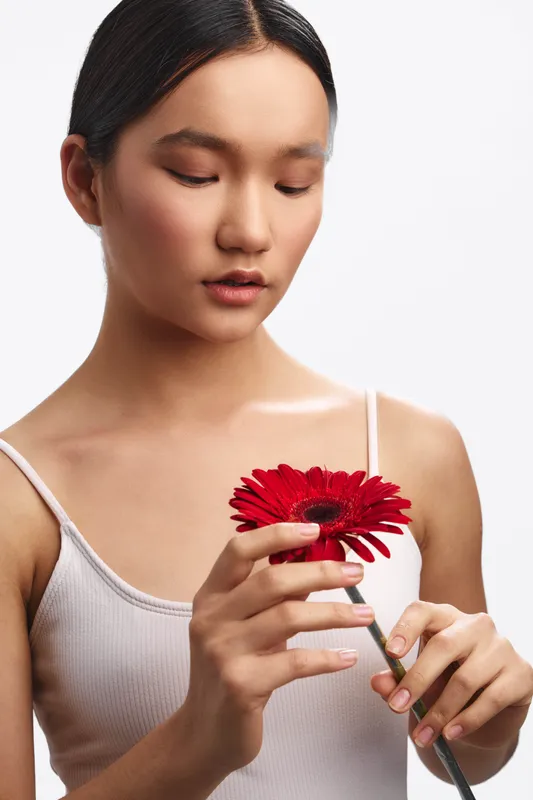 Asian woman is shown holding a vibrant red flower in a minimalist studio setting. She gazes gently at the blossom, conveying calm focus and quiet beauty.