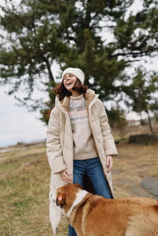 Woman outdoors with a dog in winter, wearing a coat and beanie, smiling in a park setting. Cozy casual fashion and friendly companionship in daylight.