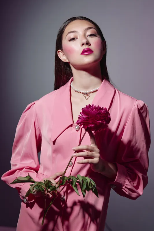 Confident young woman wears stylish pink jacket holding a dark pink flower, her bright makeup and gleaming lips emphasize beauty and fashion in creative studio portrait.
