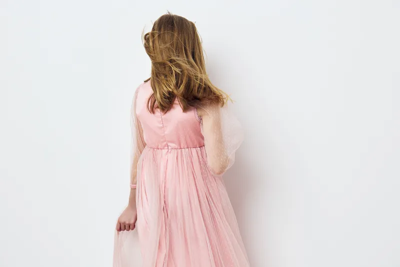 A graceful young girl wearing a pink tulle dress, with flowing hair, facing away on a minimalist white background, embodying innocence and joy