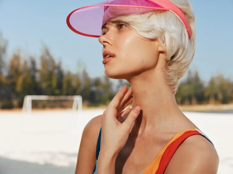 Close-up profile of a stylish woman wearing a pink visor on a sunny beach, sunlit skin, wind swept hair, vibrant colors, casual summer fashion, relaxed carefree mood.