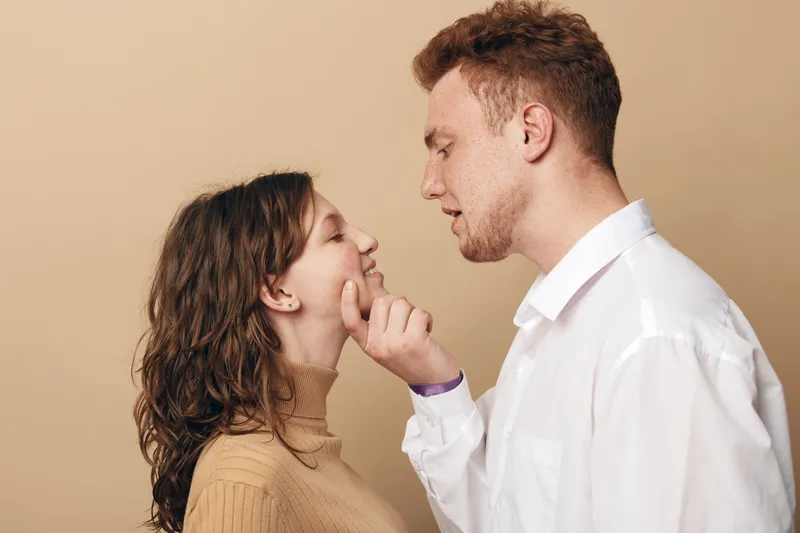 Young couple in casual clothes share intimate moment, man gently touches woman’s face. Warm beige background creates cozy romantic atmosphere and connection between partners.