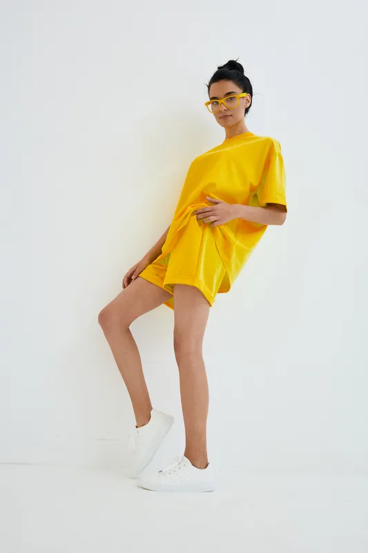 slender woman in yellow outfit poses confidently against white background, wearing oversized t-shirt, shorts, white sneakers, and yellow sunglasses, showcasing summer fashion style.