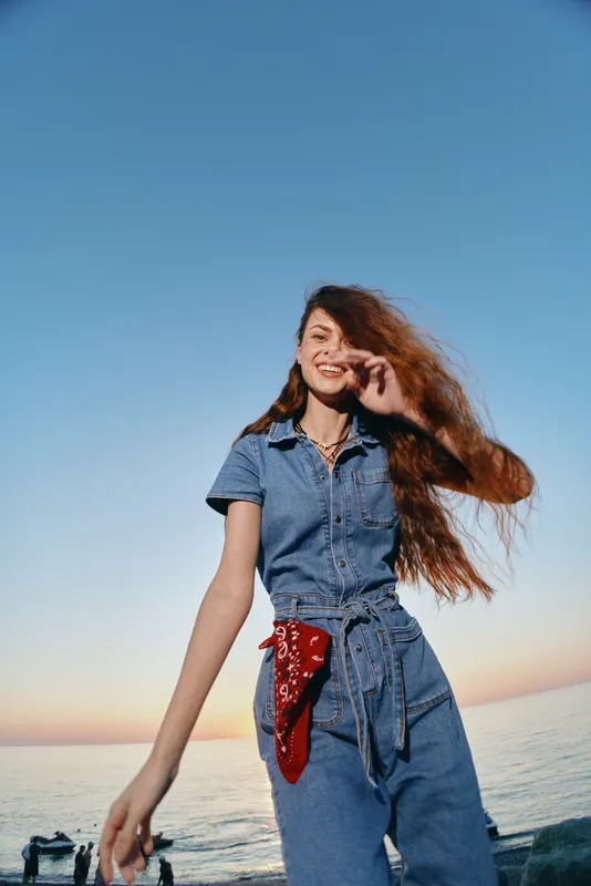 Lifestyle woman in modern boho-western denim street style with a red bandana by the beach under warm natural light and film color tones, smiling and carefree.