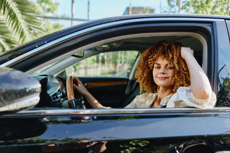 Smiling woman with curly hair sitting inside a black car, wearing a beige shirt, enjoying a sunny day with green trees in the background and a relaxed expression.