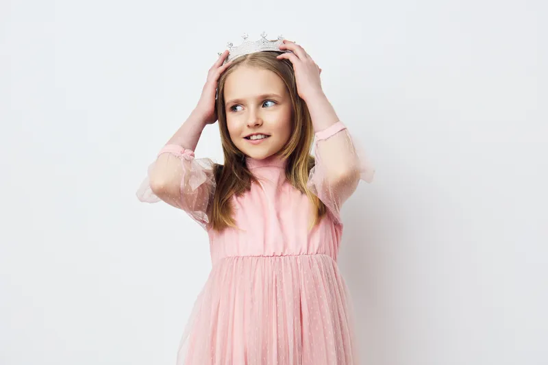 A joyful young girl in a pink dress with a crown, expressing delight and playfulness against a soft white background, ideal for children related themes