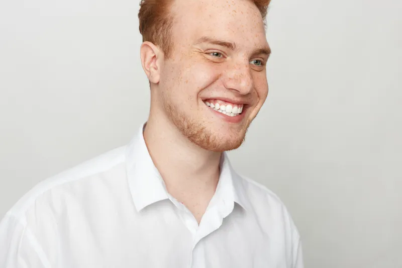 Smiling young man with red hair and freckles wears white shirt, looking away. Bright studio portrait with soft lighting, concept of happiness, youth, and positive emotions.