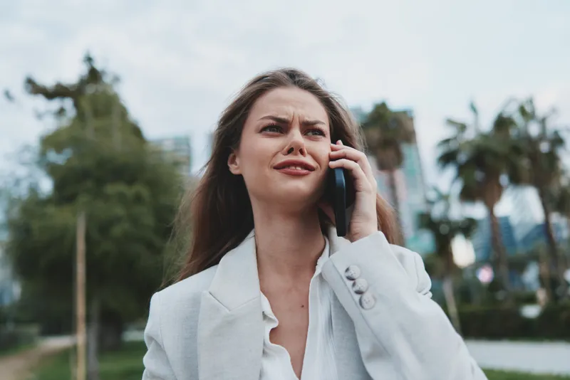 Stressed woman in white blazer talking on phone with worried expression outdoors urban background financial crisis concept