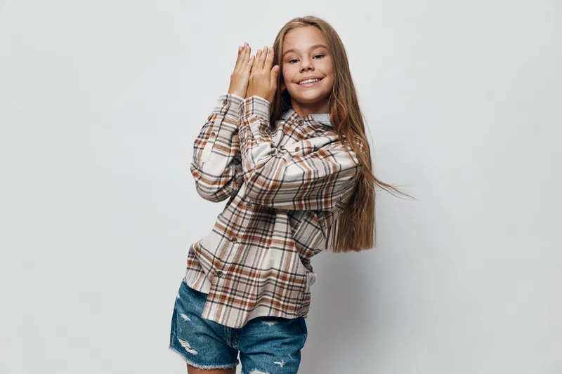 Girl with long hair smiling and posing with crossed hands wearing plaid shirt and jeans shorts on light gray background in studio. Child casual clothing and happiness.