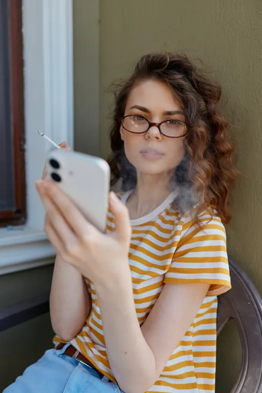 Selfie taking young woman with curly hair sits by a window in a striped tee, holding a phone and gazing at the screen with a calm, focused expression, creating a natural home moment.