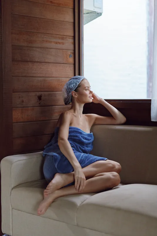 Relaxed woman sitting on sofa wrapped in blue towel with hair wrapped in shower cap, enjoying natural light from window in cozy wooden room interior.