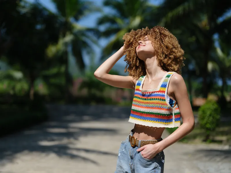 young woman with curly hair wearing colorful crochet top and denim shorts standing outdoors, enjoying sunny weather, smiling with eyes closed, tropical background
