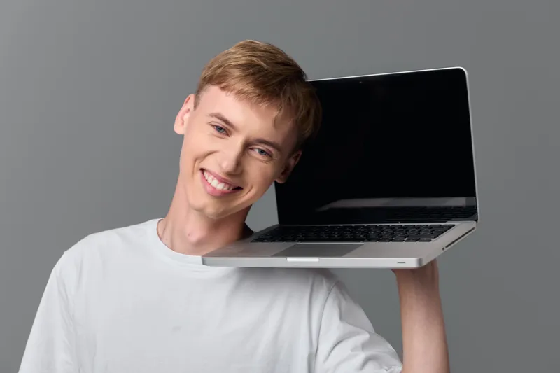 Smiling man balances a laptop on his shoulder in a casual studio setup, highlighting technology, multitasking, and modern workflow in a dynamic work scene