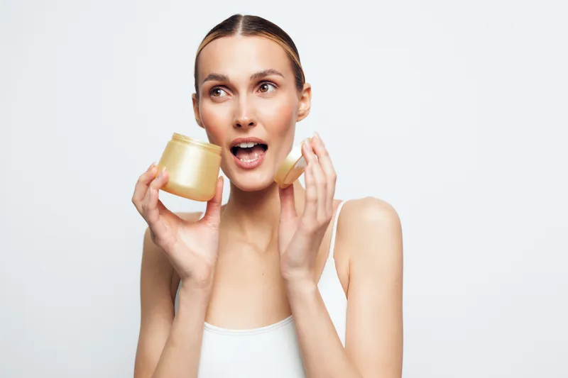 Young woman with clean skin holding an open jar of cream applying moisturizer on her face, natural beauty concept, white background, skincare routine, fresh look