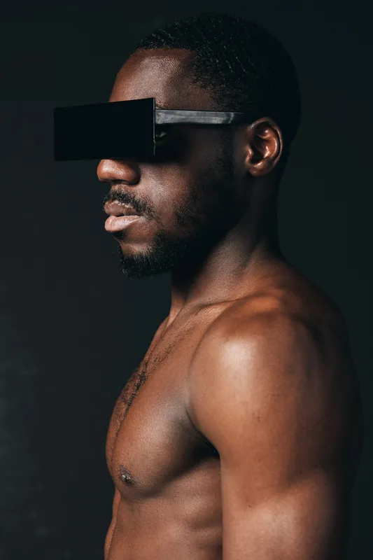 Creative conceptual portrait of African man with dark skin wearing futuristic black glasses in visual style, isolated on dark background with strong lighting and shadow contrast.