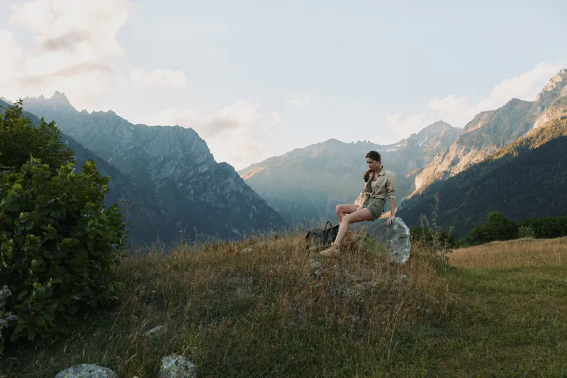 A woman sits on a rock in a mountain meadow, enjoying the view, calm and reflective. A nature driven moment that conveys outdoor lifestyle and solo exploration.
