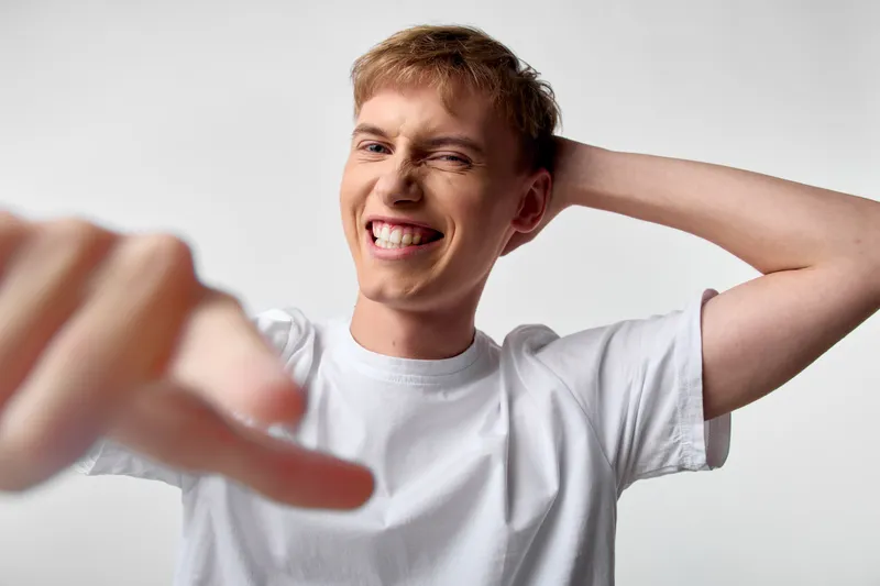 Joyful young man in a white T shirt smiles widely while pointing toward the camera, capturing a dynamic, playful moment of confidence and energy