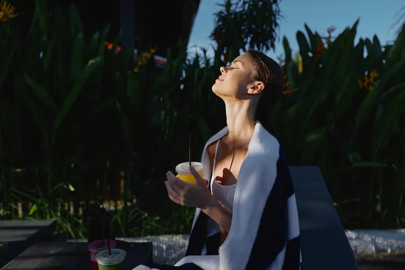 Outdoor sun relaxation and leisure in a garden setting, a person with a towel draped over shoulders enjoys a beverage, creating a casual lifestyle moment of summer calm and well being.