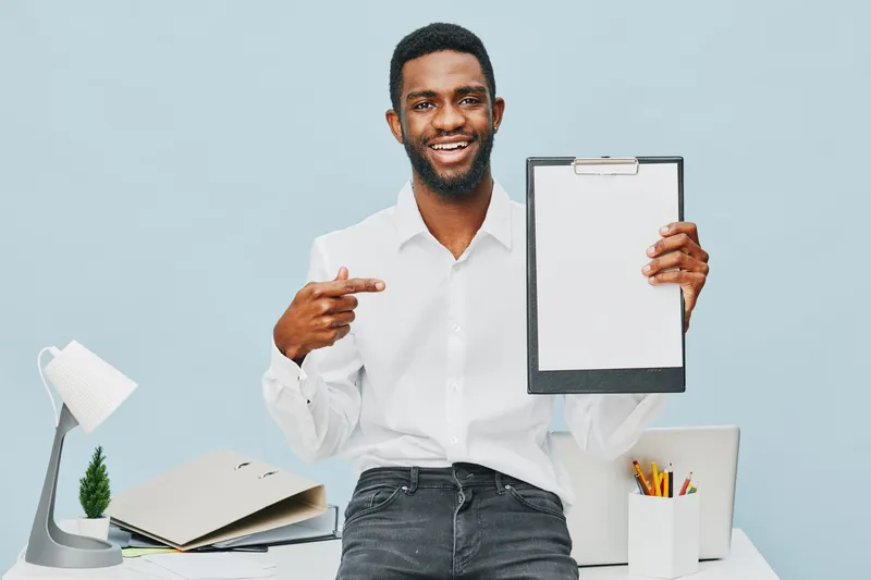 Smiling man in white shirt holding clipboard with blank sheet in office workspace studio shot. Happy professional young African American male presenting document for business or presentation concept.