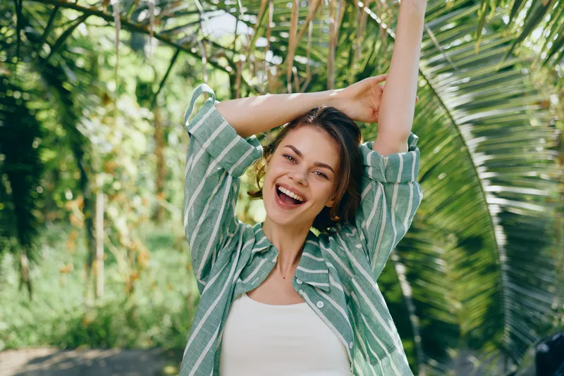 Brightly lit portrait of a joyful woman outdoors, arms raised, broad smile, casual striped shirt, sunny natural backdrop.