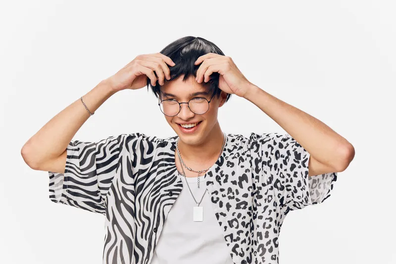 Smiling young man wearing glasses and a patterned shirt with zebra and leopard prints poses cheerfully against a white background, showing playful energy and style.