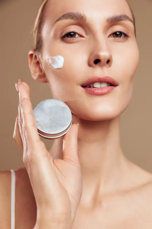 Woman skincare portrait, cosmetic beauty close up, face cream on cheek, holding compact mirror, hand gesture, calm expression, studio lighting, smooth skin, beige background, soft natural glow