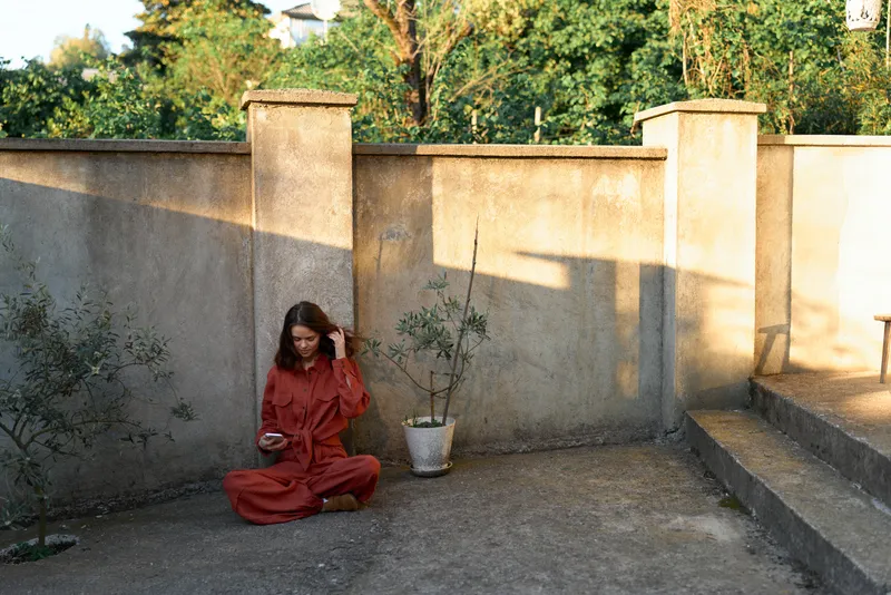 A contemplative woman in a red dress sits on the ground beside a stone wall, outdoors in a quiet courtyard with sunlight and greenery