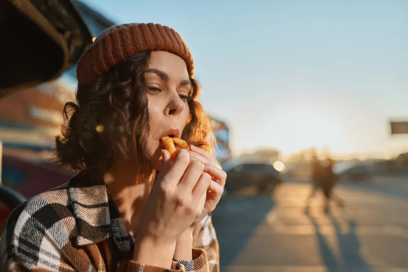 Woman eating fries on an urban street at golden hour, portrait with beanie and warm sunlight, candid lifestyle moment of authenticity, mindful living and emotional storytelling in a cozy scene