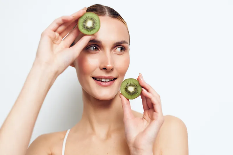 A smiling young woman holds two halves of a fresh kiwi fruit near her face against a white background. The concept highlights healthy lifestyle and natural beauty with fresh fruit.