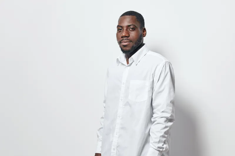 African American man with dark skin wearing white shirt standing against white background looking at camera with calm expression and relaxed posture in studio lighting.