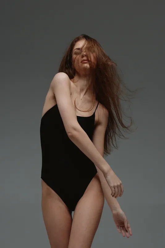 Red-haired woman in black bodysuit posing gracefully with flowing hair and neutral gray background.