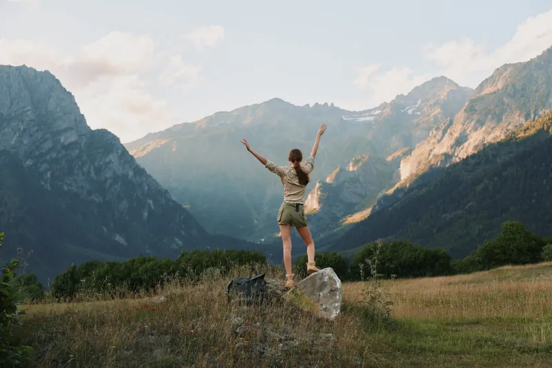 A woman in casual clothes stands on a rock with arms raised, facing mountain peaks under a partly cloudy sky. The scene captures freedom and outdoor lifestyle.
