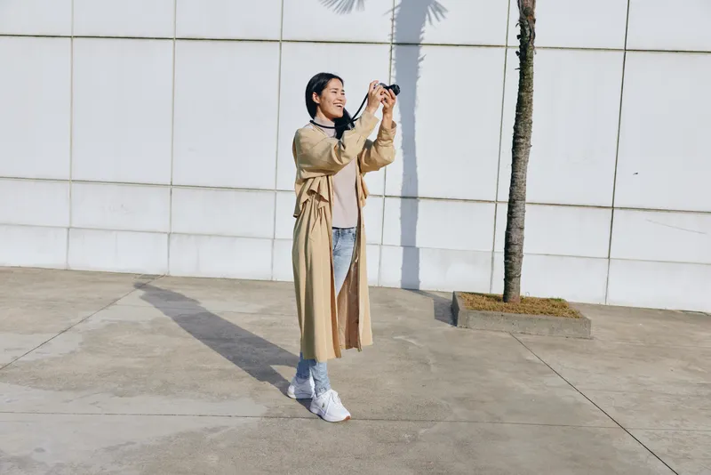 Photography, woman, outdoor, camera, street, fashion, trenchcoat, jeans, sneakers, urban, daylight, lifestyle, casual, model, candid, streetwear, clothing, coat, jacket, sunlight, pavement, sidewalk,