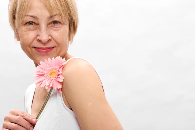 Woman smile closeup portrait with pink flower held to shoulder, elderly blonde hair, soft studio light on white background, happiness, beauty, calm mood