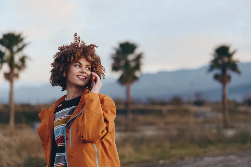 Smiling woman in orange jacket outdoors, curly hair blowing gently, holding phone to ear, relaxed lifestyle moment, golden hour light, open field with palms and distant mountains