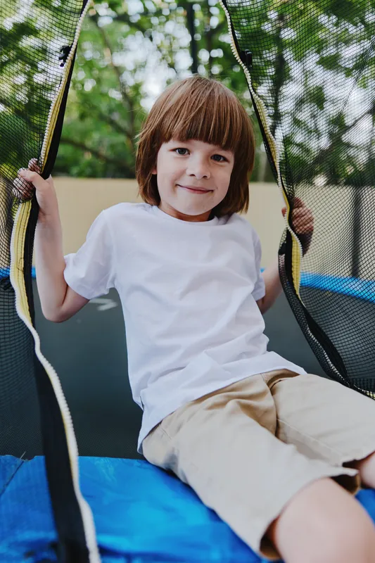 Happy boy playing on a trampoline in a sunny outdoor setting, showcasing joy and excitement in a playful moment, surrounded by lush greenery.