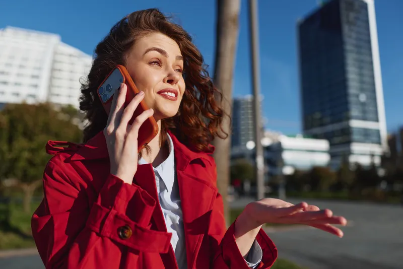 Young woman in a red trench talks on a smartphone outdoors, smiling under sunny urban surroundings, expressive and confident in a candid city moment