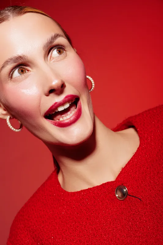 Close-up of a smiling woman with bright red lipstick and red sweater against red background. She wears hoop earrings and looks joyful. Concept of fashion, beauty, and vibrant style.