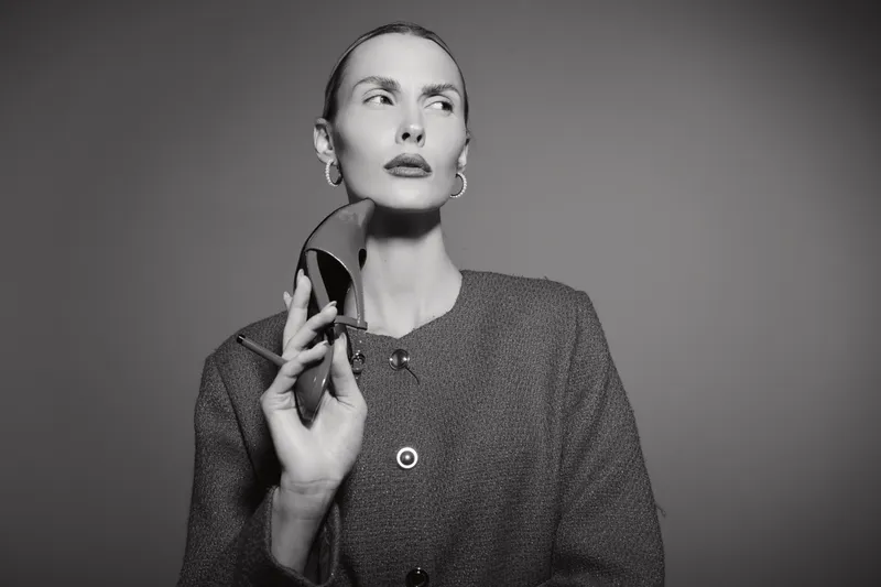 Elegant white woman with pale skin and slicked back hair holds high heel shoe near face, wearing textured jacket. Studio portrait in black and white with dramatic lighting.