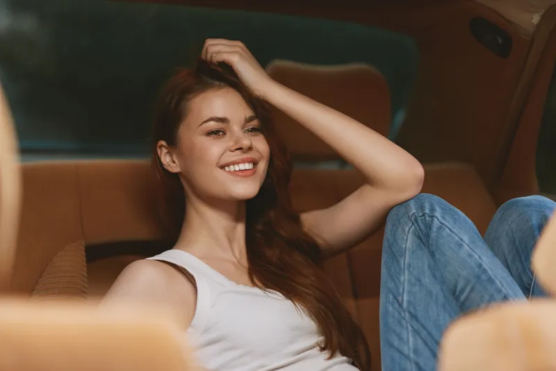 Young woman smiling in a car, wearing a white tank top and blue jeans, with a relaxed pose and warm lighting, conveying happiness and comfort.